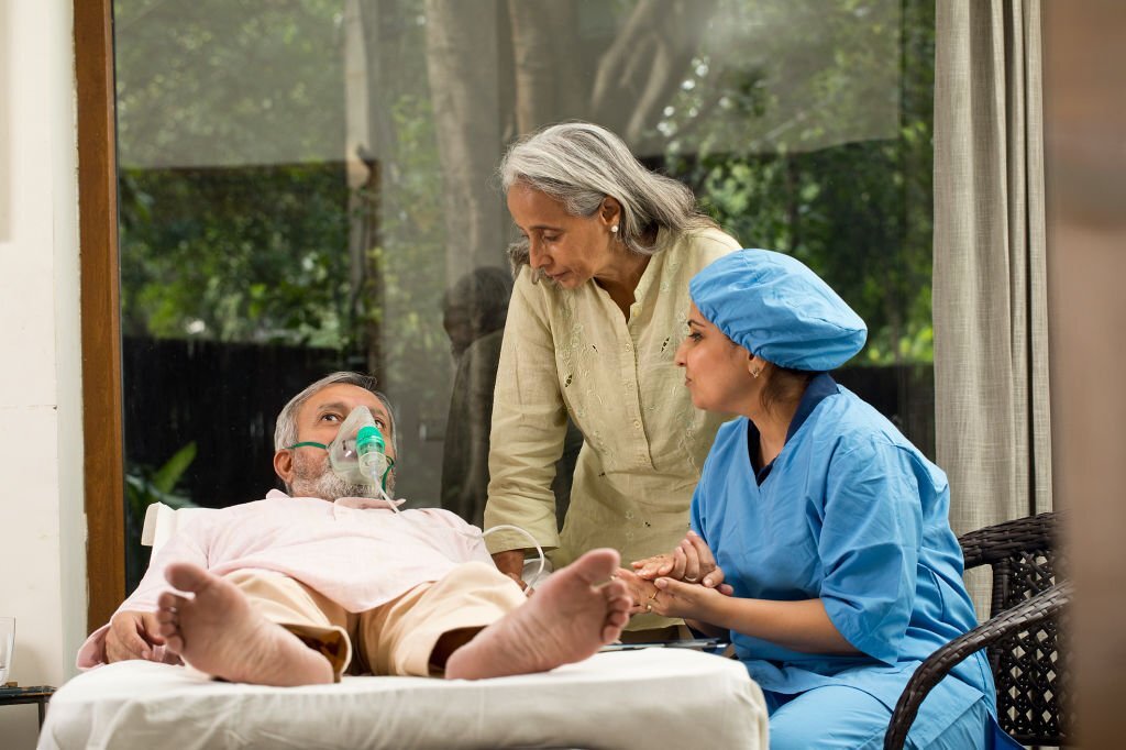 Female nurse explaining to woman while old male patient is lying on bed